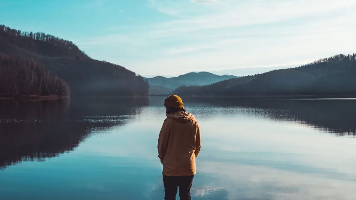 Person in Kapuzenpullover steht am Ufer eines spiegelglatten Bergsees