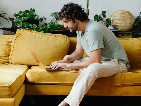 Ein Mann sitzt entspannt auf einem gelben Sofa und tippt auf seinem Laptop, im Hintergrund stehen Zimmerpflanzen.